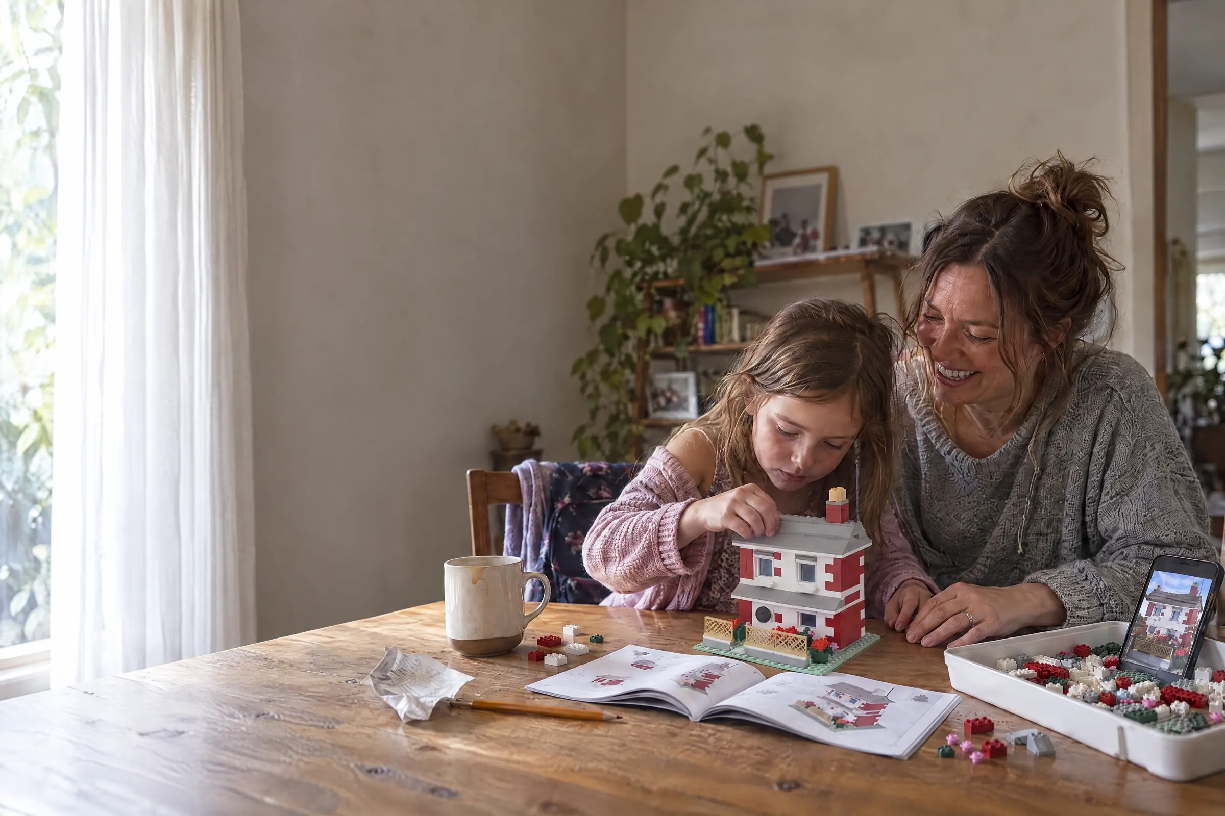 A mother and daughter build a brick model of their house at the dining table, with the instruction booklet open and a photo of the real house on a phone